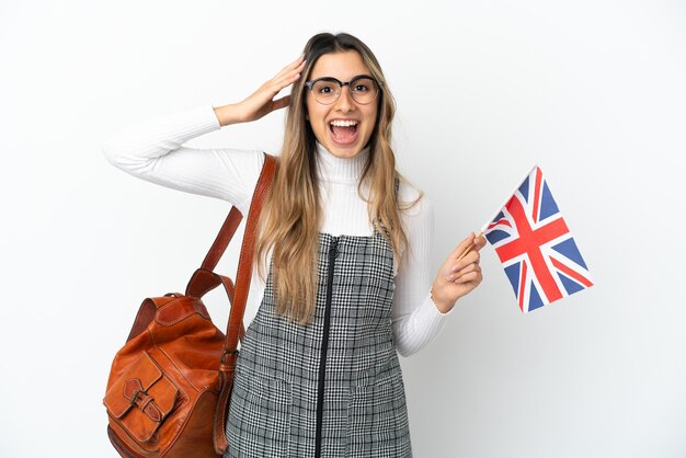 Jonge kaukasische vrouw die een vlag van het Verenigd Koninkrijk houdt die op witte achtergrond met verrassingsuitdrukking wordt geïsoleerd