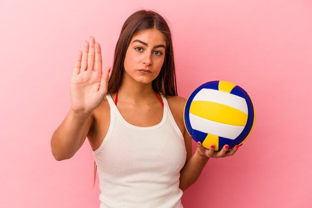 Jonge kaukasische vrouw die een volleybalbal houdt die op roze achtergrond wordt geïsoleerd die zich met uitgestrekte hand bevindt die stopbord toont, die u verhinderen.