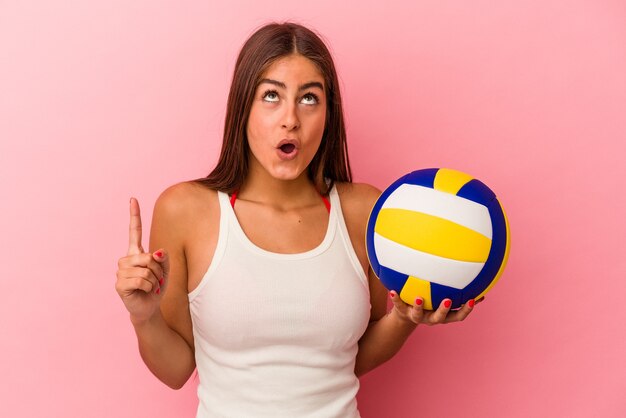 Jonge kaukasische vrouw die een volleyballbal houdt die op roze achtergrond wordt geïsoleerd die omhoog wijst met geopende mond.