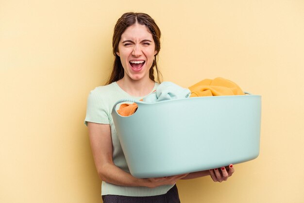 Jonge kaukasische vrouw die een wasmand houdt die op gele achtergrond wordt geïsoleerd en erg boos en agressief schreeuwt