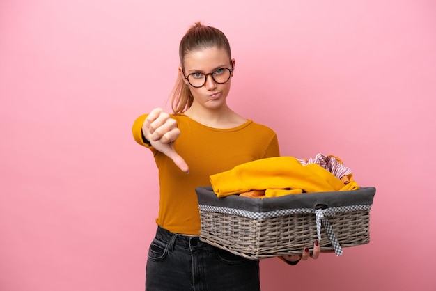 Jonge kaukasische vrouw die een wasmand houdt die op roze achtergrond wordt geïsoleerd die duim omlaag met negatieve uitdrukking toont