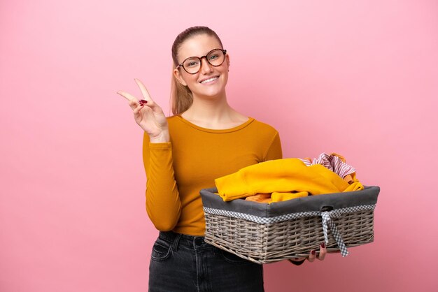 Jonge kaukasische vrouw die een wasmand houdt die op roze achtergrond wordt geïsoleerd en glimlacht en overwinningsteken toont