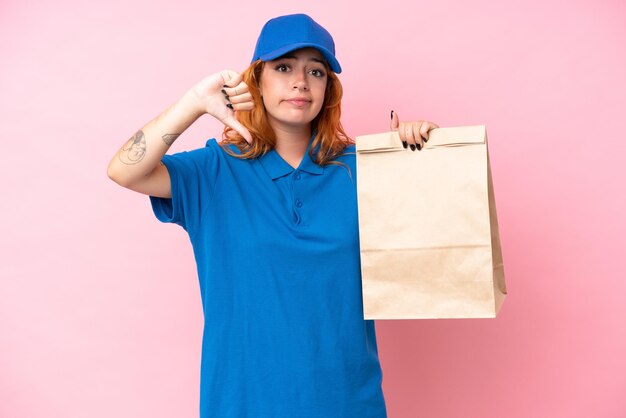 Jonge kaukasische vrouw die een zak afhaalmaaltijden neemt die op roze achtergrond wordt geïsoleerd die duim neer met negatieve uitdrukking toont