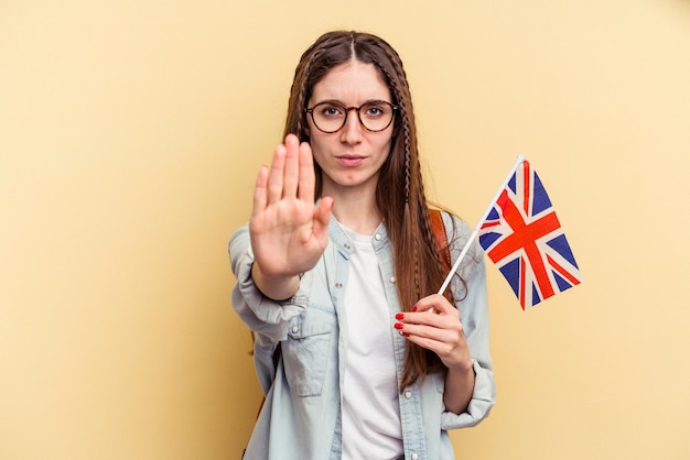 Jonge kaukasische vrouw die Engels studeert geïsoleerd op een gele achtergrond die staat met uitgestrekte hand met stopbord dat je verhindert