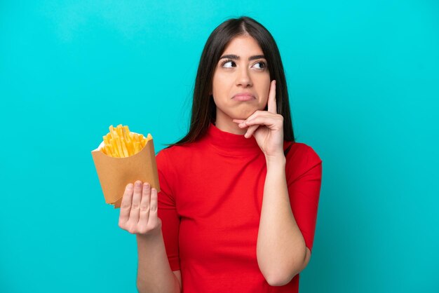 Jonge kaukasische vrouw die frietjes vangt die op blauwe achtergrond worden geïsoleerd en een idee denkt