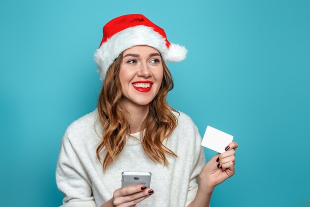 Jonge kaukasische vrouw die in witte sweater en santahoed creditcard en mobiele telefoon houdt die aan de kant kijken en glimlachen geïsoleerd op blauwe muur