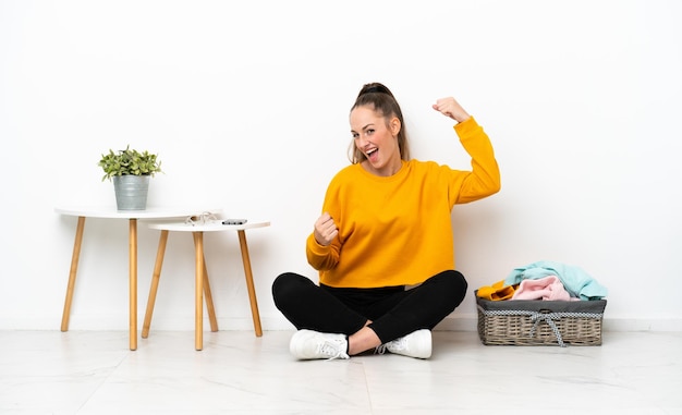 Jonge kaukasische vrouw die kleren vouwt die op de vloer zitten die op witte achtergrond wordt geïsoleerd en een overwinning viert