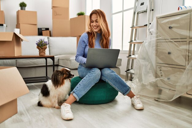 Jonge kaukasische vrouw die laptop gebruikt die met hond bij nieuw huis zit