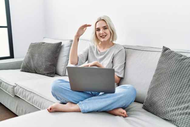 Jonge kaukasische vrouw die laptop thuis gebruiken zittend op de bank gebarend met handen die groot en groot tekenmaatregelsymbool tonen glimlachend kijkend naar het meetconcept van de camera