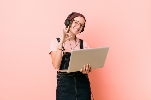 Jonge Kaukasische vrouw die laptop vrolijk en zeker houdt die ok gebaar toont.