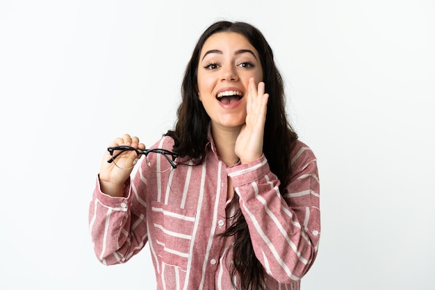 Jonge kaukasische vrouw die met glazen op witte achtergrond wordt geïsoleerd die met wijd open mond schreeuwen