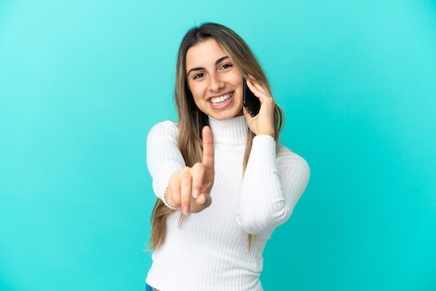 Jonge kaukasische vrouw die mobiele telefoon gebruikt die op blauwe achtergrond wordt geïsoleerd en een vinger toont en opheft