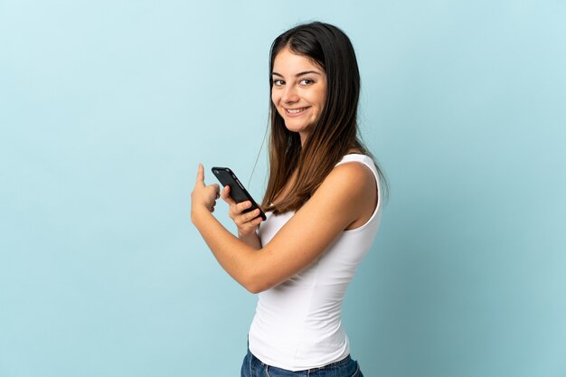 Jonge kaukasische vrouw die mobiele telefoon met behulp van die op blauw wordt geïsoleerd