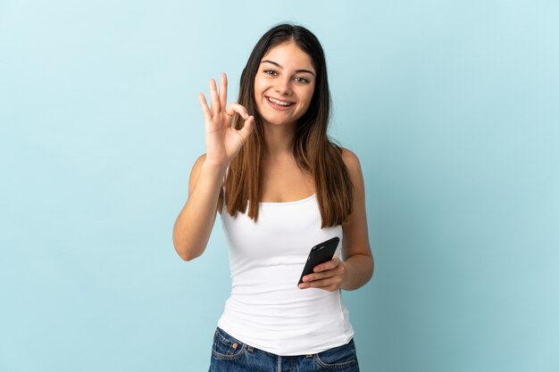 Jonge kaukasische vrouw die mobiele telefoon met behulp van die op blauwe achtergrond wordt geïsoleerd die ok teken met vingers toont