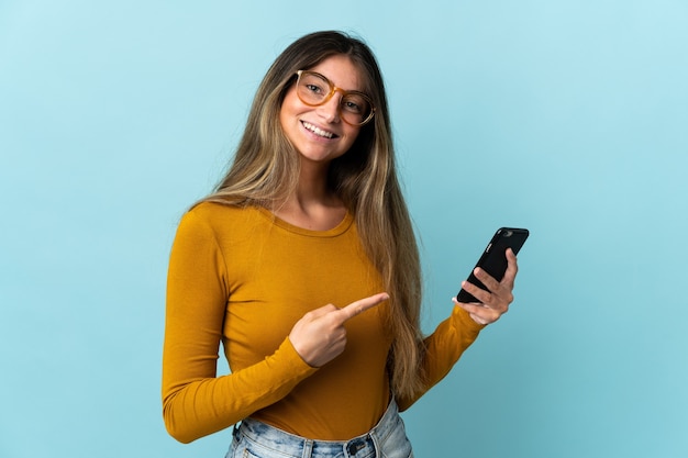 Jonge kaukasische vrouw die mobiele telefoon met behulp van die op blauwe muur wordt geïsoleerd en het richt