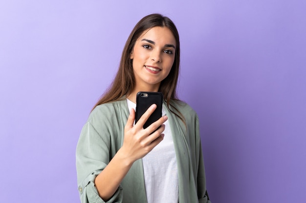 Jonge kaukasische vrouw die mobiele telefoon met behulp van die op purpere muur met gelukkige uitdrukking wordt geïsoleerd