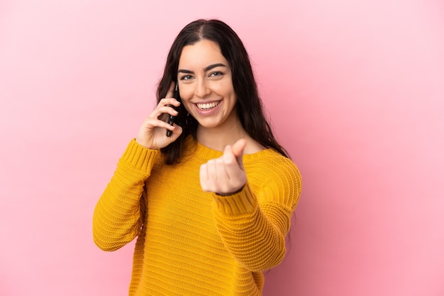 Jonge kaukasische vrouw die mobiele telefoon met behulp van die op roze achtergrond wordt geïsoleerd die geldgebaar maakt