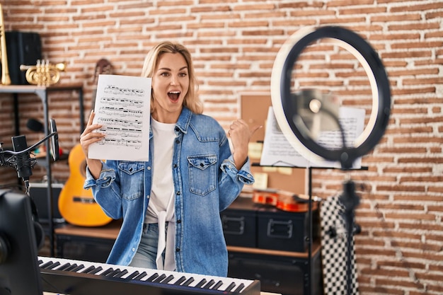 Jonge kaukasische vrouw die online muziekleerprogramma doet die muziekblad tonen die duim omhoog aan de kant richten die gelukkig met open mond glimlachen