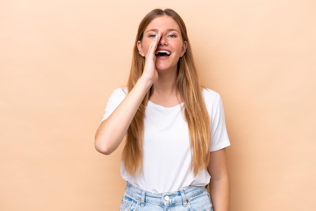 Jonge kaukasische vrouw die op beige achtergrond wordt geïsoleerd die met wijd open mond schreeuwt