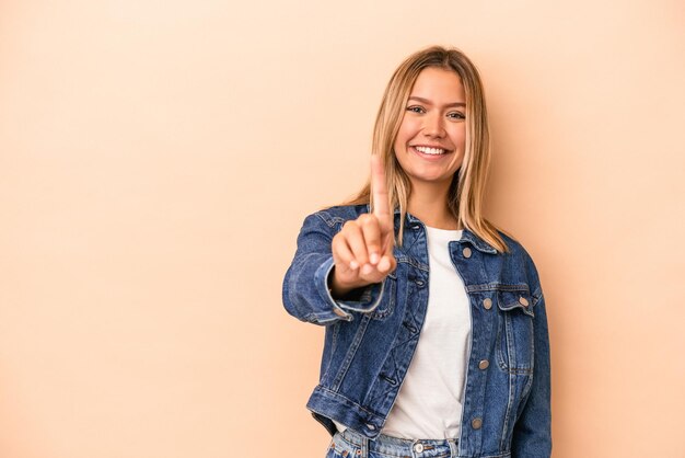 Jonge kaukasische vrouw die op beige achtergrond wordt geïsoleerd die nummer één met vinger toont.