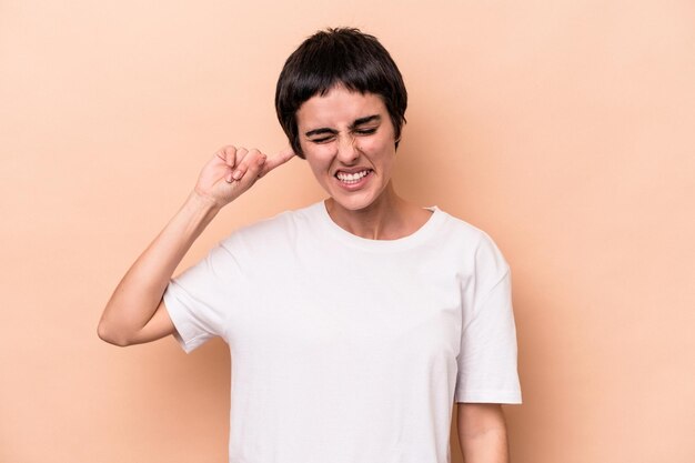 Jonge kaukasische vrouw die op beige achtergrond wordt geïsoleerd die oren behandelt met handen.