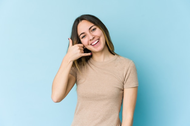 Jonge kaukasische vrouw die op blauw een gsm-gebaar met vingers toont.