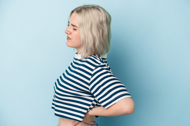 Jonge kaukasische vrouw die op blauwe achtergrond wordt geïsoleerd die aan rugpijn lijdt.