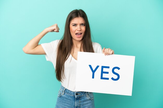 Jonge kaukasische vrouw die op blauwe muur wordt geïsoleerd die een aanplakbiljet met tekst YES houdt die sterk gebaar doet