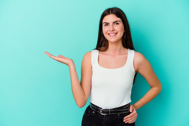 Jonge kaukasische vrouw die op blauwe muur wordt geïsoleerd die een exemplaarruimte op een palm toont en een andere hand op taille houdt.