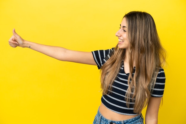 Jonge kaukasische vrouw die op gele achtergrond wordt geïsoleerd die een duim omhoog gebaar geeft