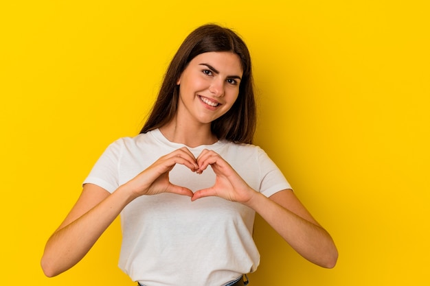 Jonge kaukasische vrouw die op gele muur wordt geïsoleerd die en een hartvorm met handen glimlacht toont.