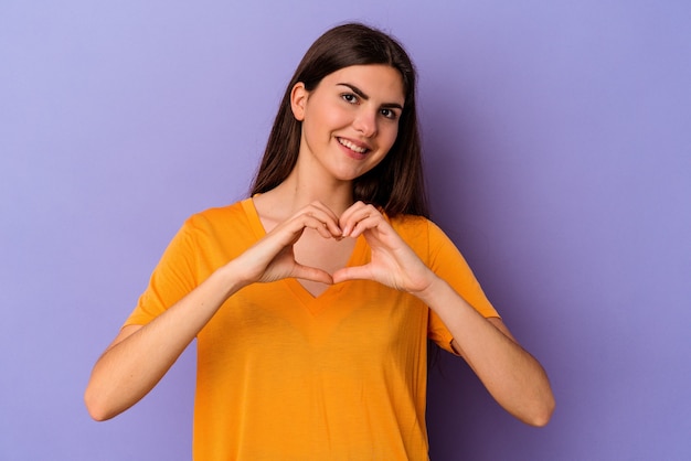 Jonge kaukasische vrouw die op purpere achtergrond wordt geïsoleerd die en een hartvorm met handen glimlacht toont.