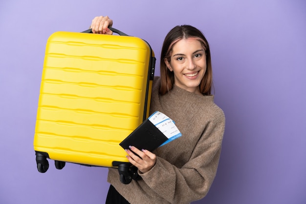 Jonge kaukasische vrouw die op purpere muur in vakantie met koffer en paspoort wordt geïsoleerd