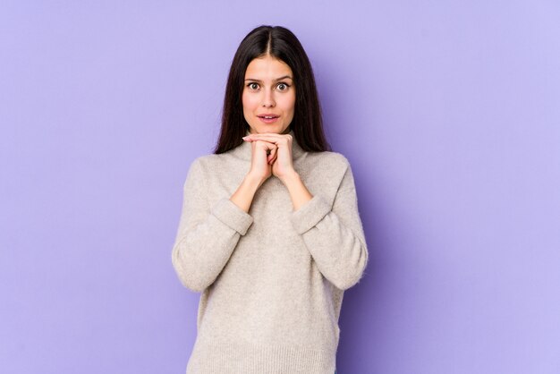 Jonge kaukasische vrouw die op purpere muur voor geluk bidden, verbaasd en mond openen die aan voorzijde kijken.