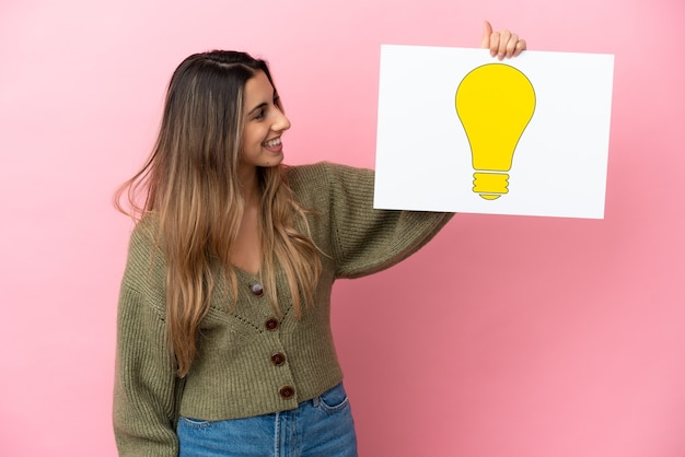 Jonge kaukasische vrouw die op roze achtergrond wordt geïsoleerd die een aanplakbiljet met bolpictogram houdt