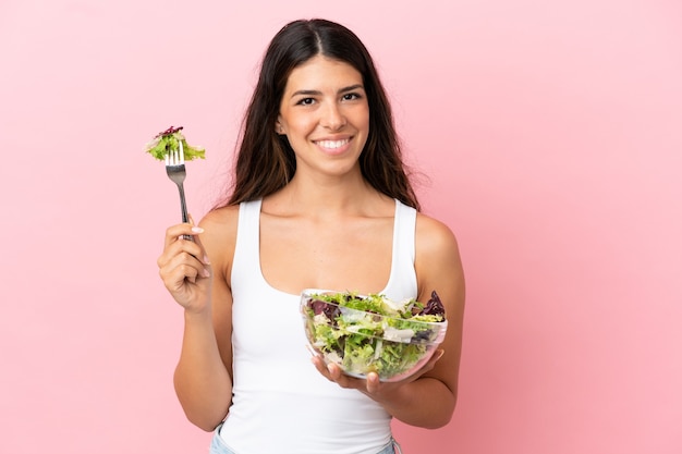 Jonge kaukasische vrouw die op roze achtergrond wordt geïsoleerd die een kom salade met gelukkige uitdrukking houdt