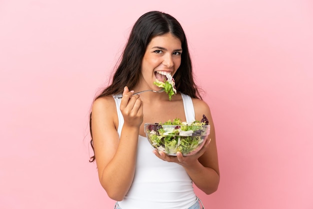 Jonge kaukasische vrouw die op roze achtergrond wordt geïsoleerd die een kom salade met gelukkige uitdrukking houdt