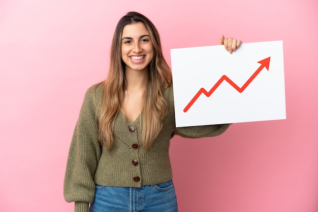 Jonge kaukasische vrouw die op roze achtergrond wordt geïsoleerd die een teken met een groeiend symbool van de statistiekenpijl met gelukkige uitdrukking houdt