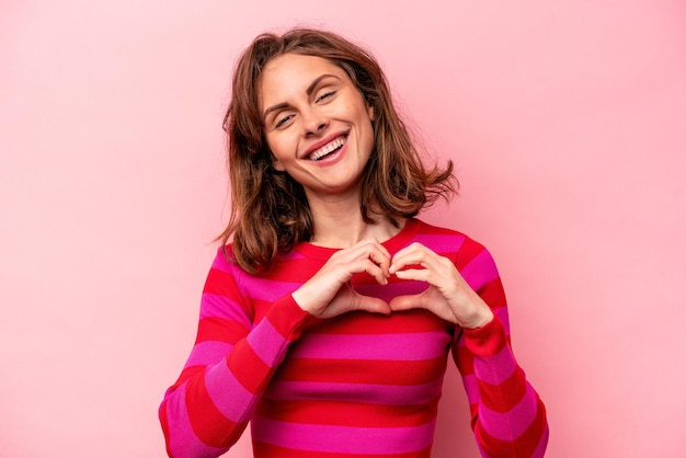 Jonge kaukasische vrouw die op roze achtergrond wordt geïsoleerd die glimlacht en een hartvorm met handen toont