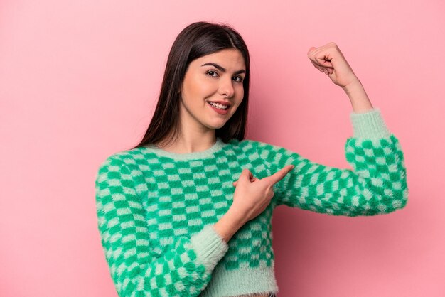 Jonge kaukasische vrouw die op roze achtergrond wordt geïsoleerd die sterktegebaar met wapenssymbool van vrouwelijke macht toont