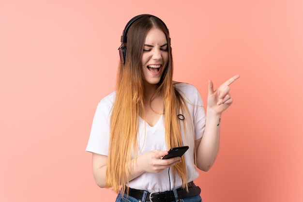 Jonge Kaukasische vrouw die op roze muur het luisteren muziek met mobiel en het zingen wordt geïsoleerd