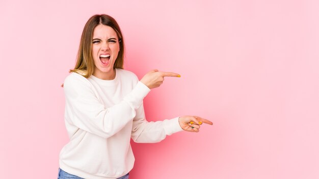 Jonge kaukasische vrouw die op roze muur wordt geïsoleerd die met wijsvingers naar een exemplaarruimte richt, opwinding en verlangen uitdrukt.