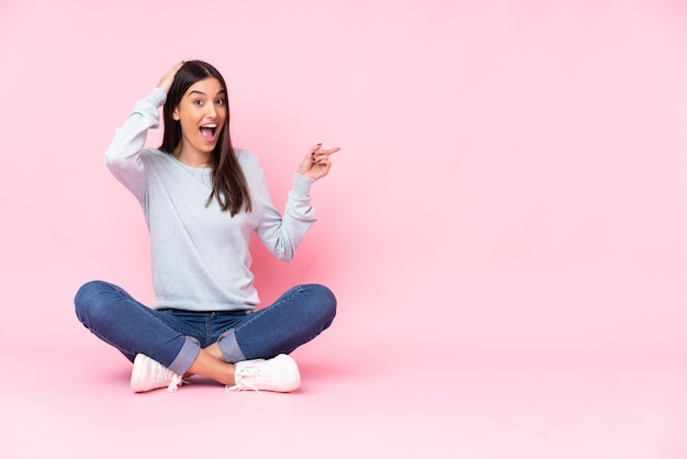 Jonge kaukasische vrouw die op roze verraste muur wordt geïsoleerd en vinger aan de kant richt