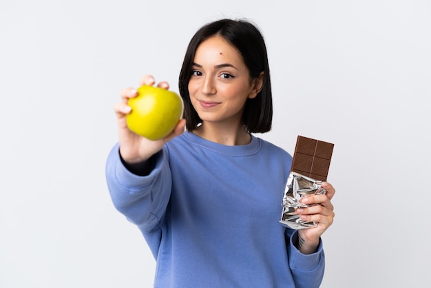 Jonge kaukasische vrouw die op witte achtergrond wordt geïsoleerd die een chocoladetablet in de ene hand en een appel in de andere neemt