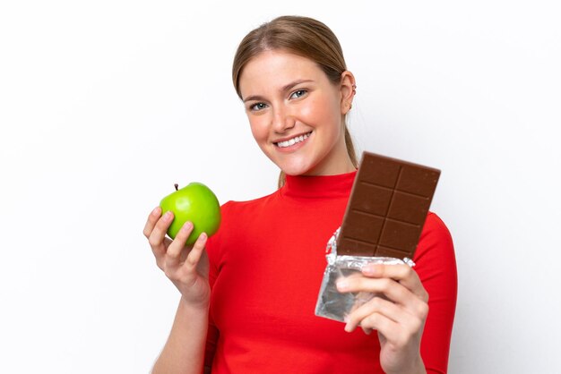 Jonge kaukasische vrouw die op witte achtergrond wordt geïsoleerd die een chocoladetablet in de ene hand en een appel in de andere neemt