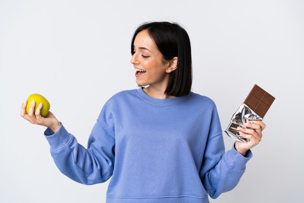 Jonge kaukasische vrouw die op witte muur wordt geïsoleerd die een chocoladetablet in één hand en een appel in andere hand neemt