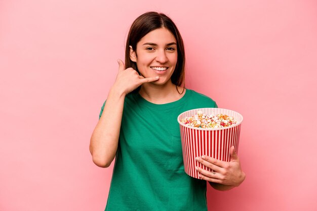 Jonge kaukasische vrouw die popcorn houdt die op roze achtergrond wordt geïsoleerd die een mobiel telefoongesprekgebaar met vingers toont