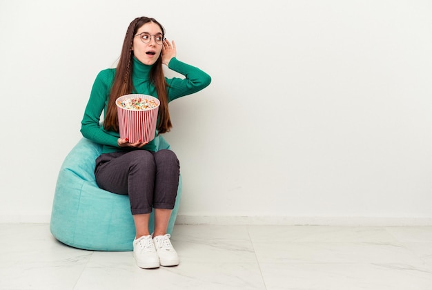 Jonge kaukasische vrouw die popcorns eet op een trekje dat op een witte achtergrond wordt geïsoleerd en probeert te luisteren naar een roddel.