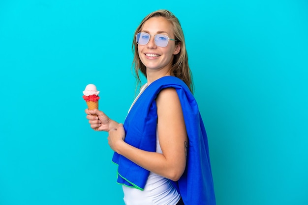 Jonge kaukasische vrouw die roomijs houdt en een strandhanddoek draagt die op blauwe achtergrond wordt geïsoleerd die veel glimlacht