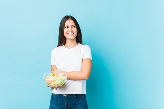 Jonge Kaukasische vrouw die salade glimlachen houden zeker met gekruiste wapens.